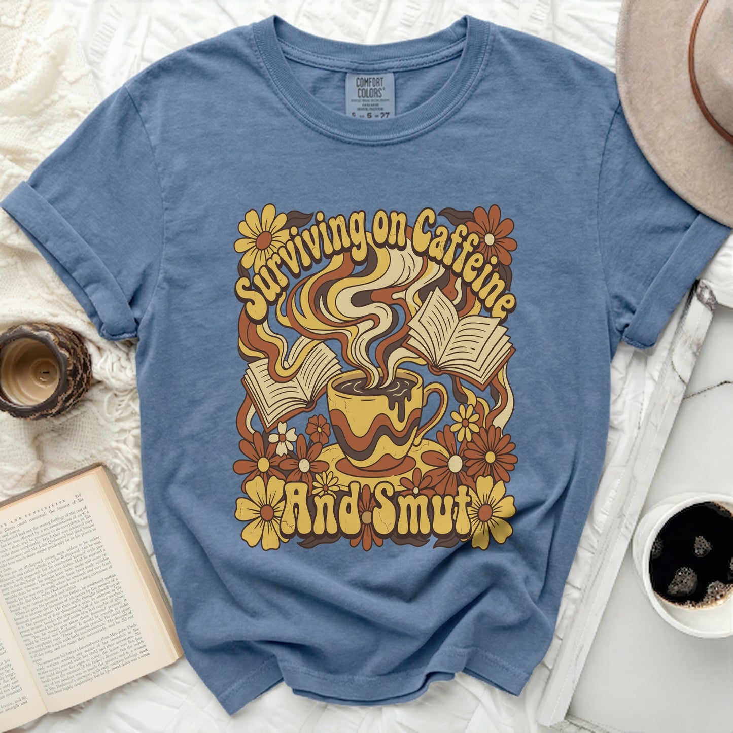 Blue t-shirt with graphic of coffee cup, books, and flowers; text reads "Surviving on Caffeine And Smut".