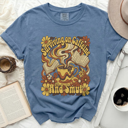 Blue t-shirt with graphic of coffee cup, books, and flowers; text reads "Surviving on Caffeine And Smut".