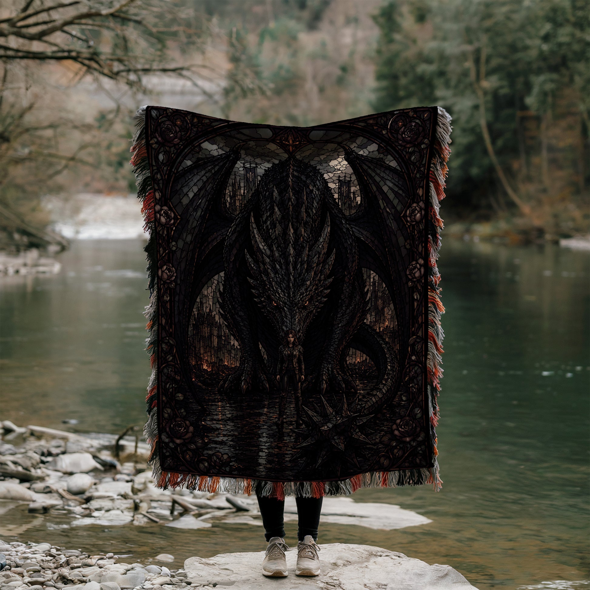 A person stands on a rock, holding a large, intricately designed dragon-themed blanket over their head, with a body of water and trees in the background.