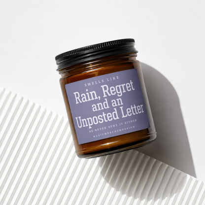 Amber glass candle with a label reading "Smells Like Rain, Regret and an Unposted Letter".