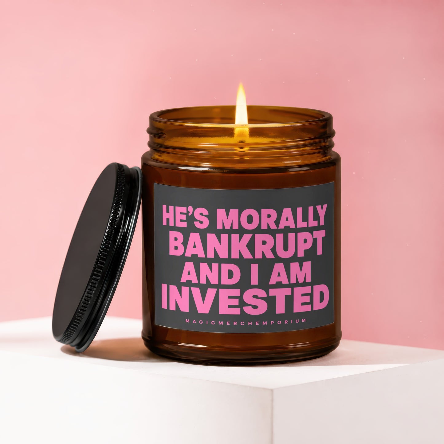 Amber candle with lid off, candle burning, text reads "HE'S MORALLY BANKRUPT AND I AM INVESTED".