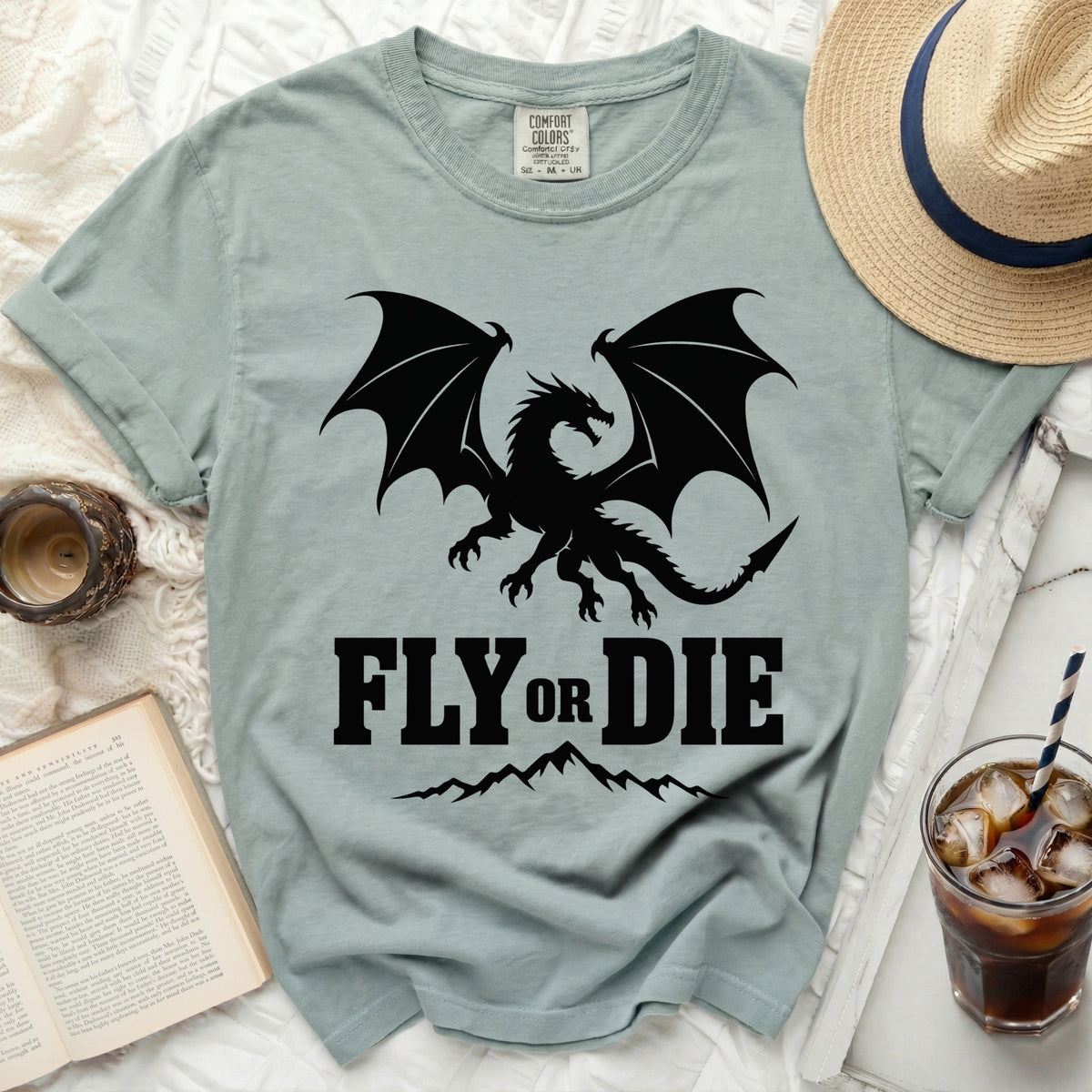 A sage green t-shirt featuring a black dragon graphic and the text FLY OR DIE, laid flat with accessories.