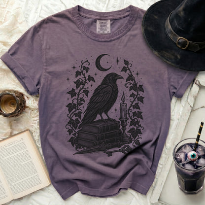 A purple t-shirt features a black graphic of a crow on books, a crescent moon, and ivy.