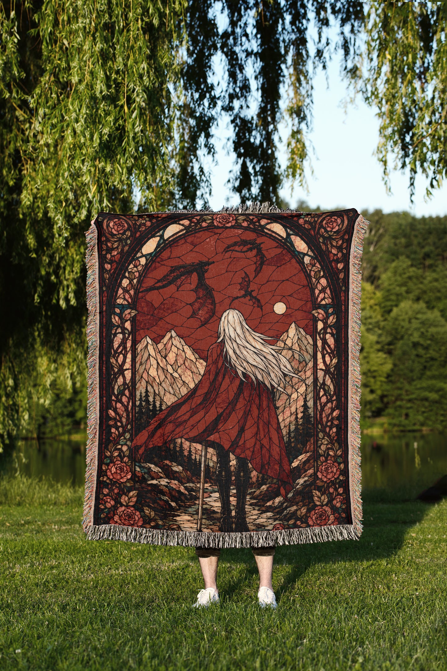 A person stands in a grassy field, holding a large, ornate rug with a red and black design depicting a woman with long hair and a flowing dress, standing in front of a scenic landscape with mountains and a lake.