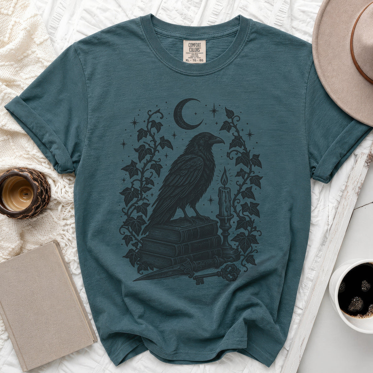 Teal t-shirt featuring a black graphic of a crow on books, framed by ivy and a crescent moon.
