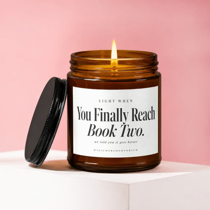 A lit amber glass jar candle with a black lid and white label against a pink background.