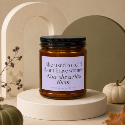 Amber candle with black lid, reading "She used to read about brave women. Now she writes them."