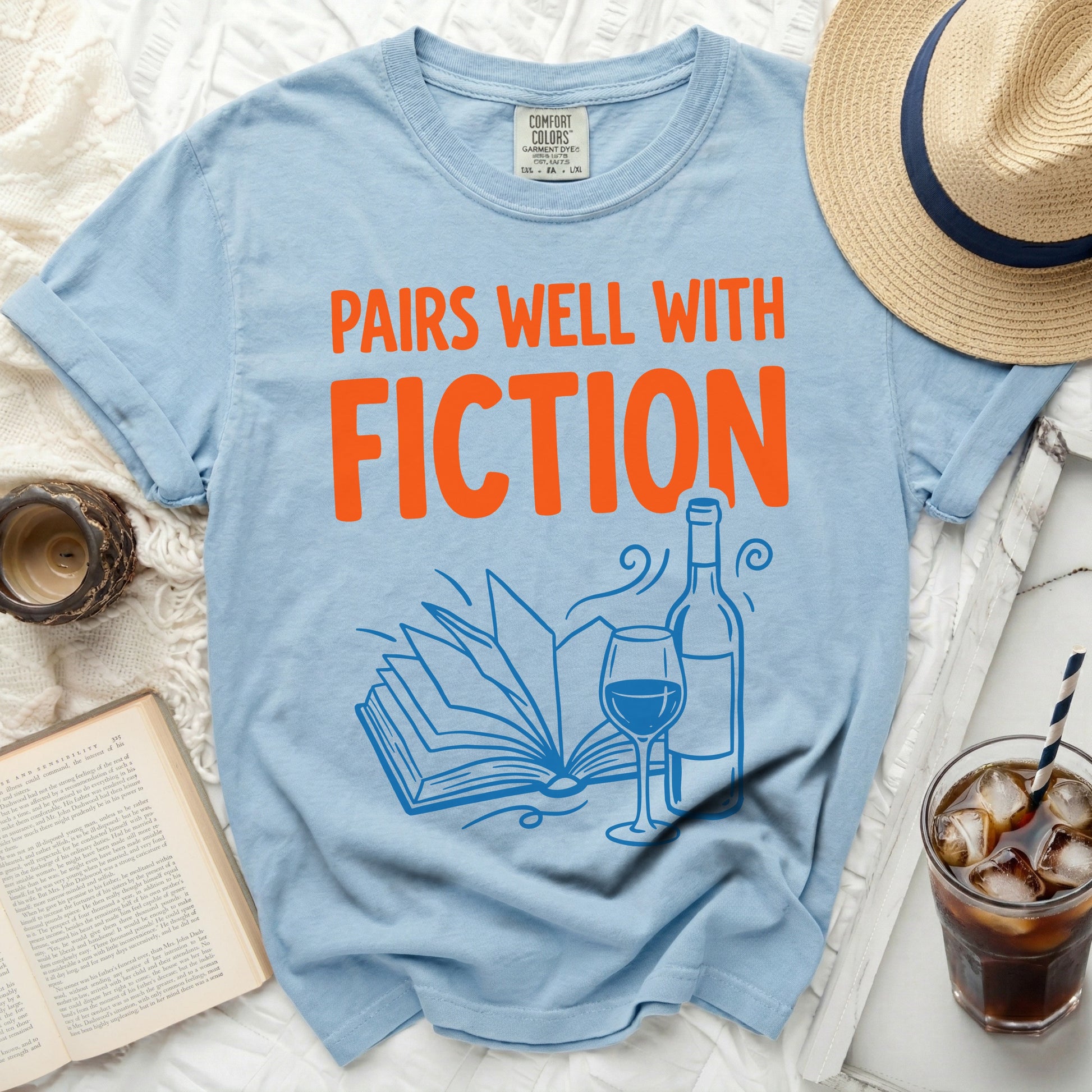 Light blue t-shirt with orange text "Pairs Well With Fiction" and an illustration of a book and wine.