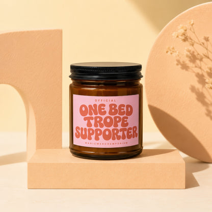 Amber glass candle with pink label reading "One Bed Trope Supporter".