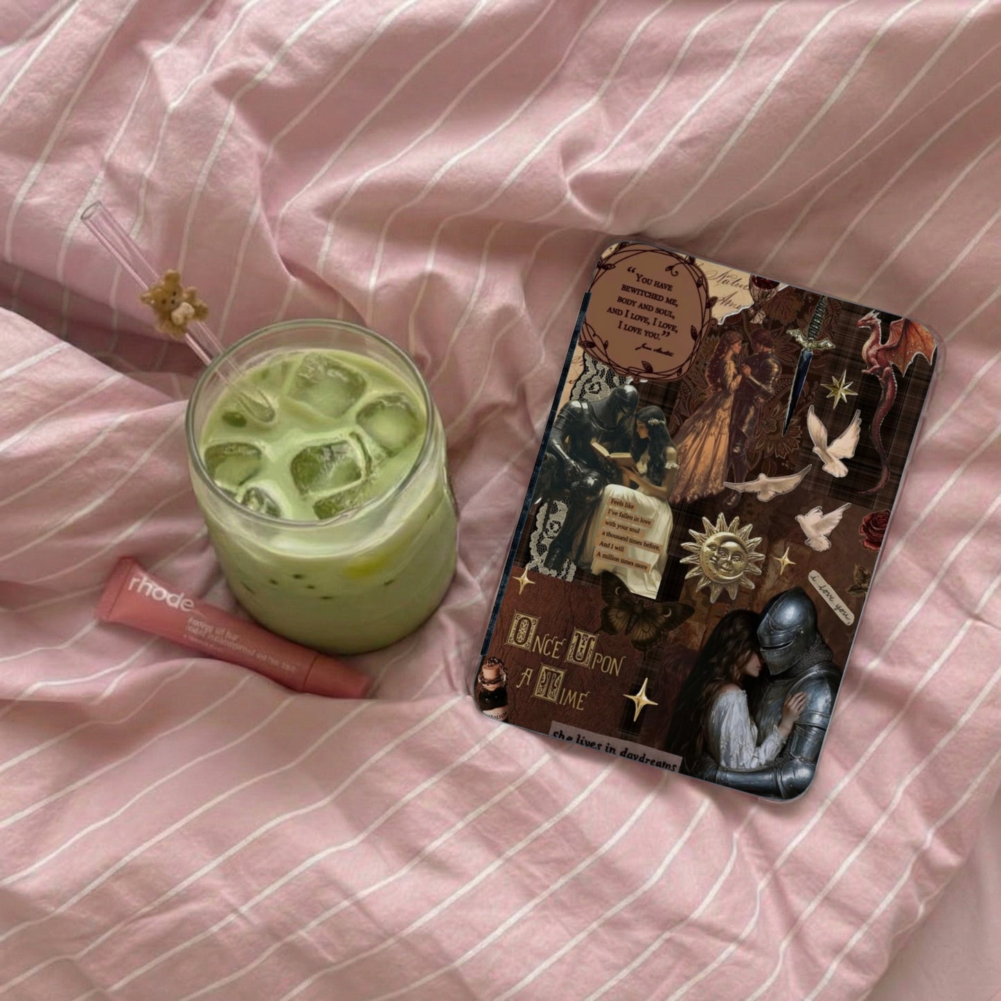A matcha drink, Rhode lip balm, and a collage art piece on pink striped fabric.