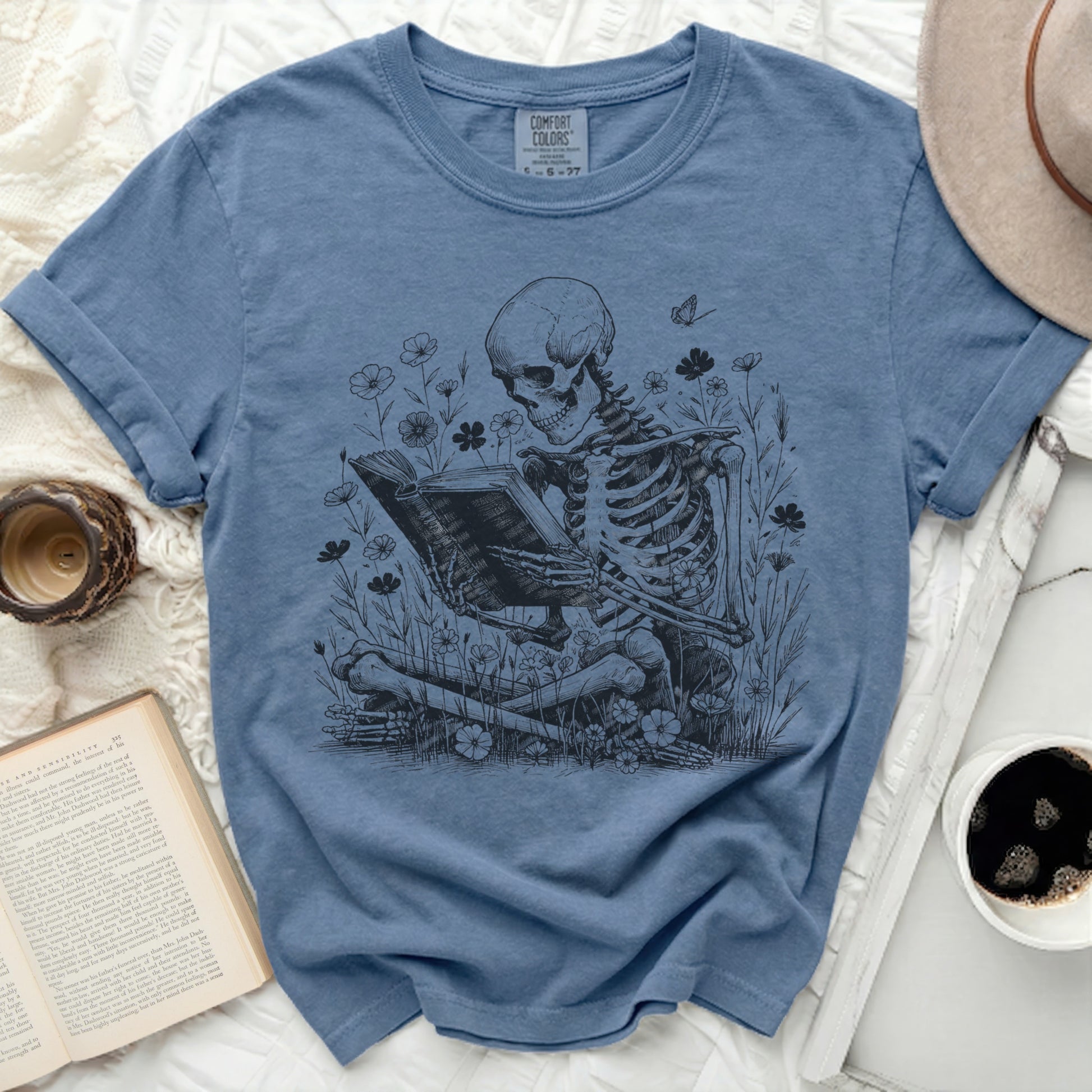 Skeleton reading a book surrounded by flowers, on a blue t-shirt.