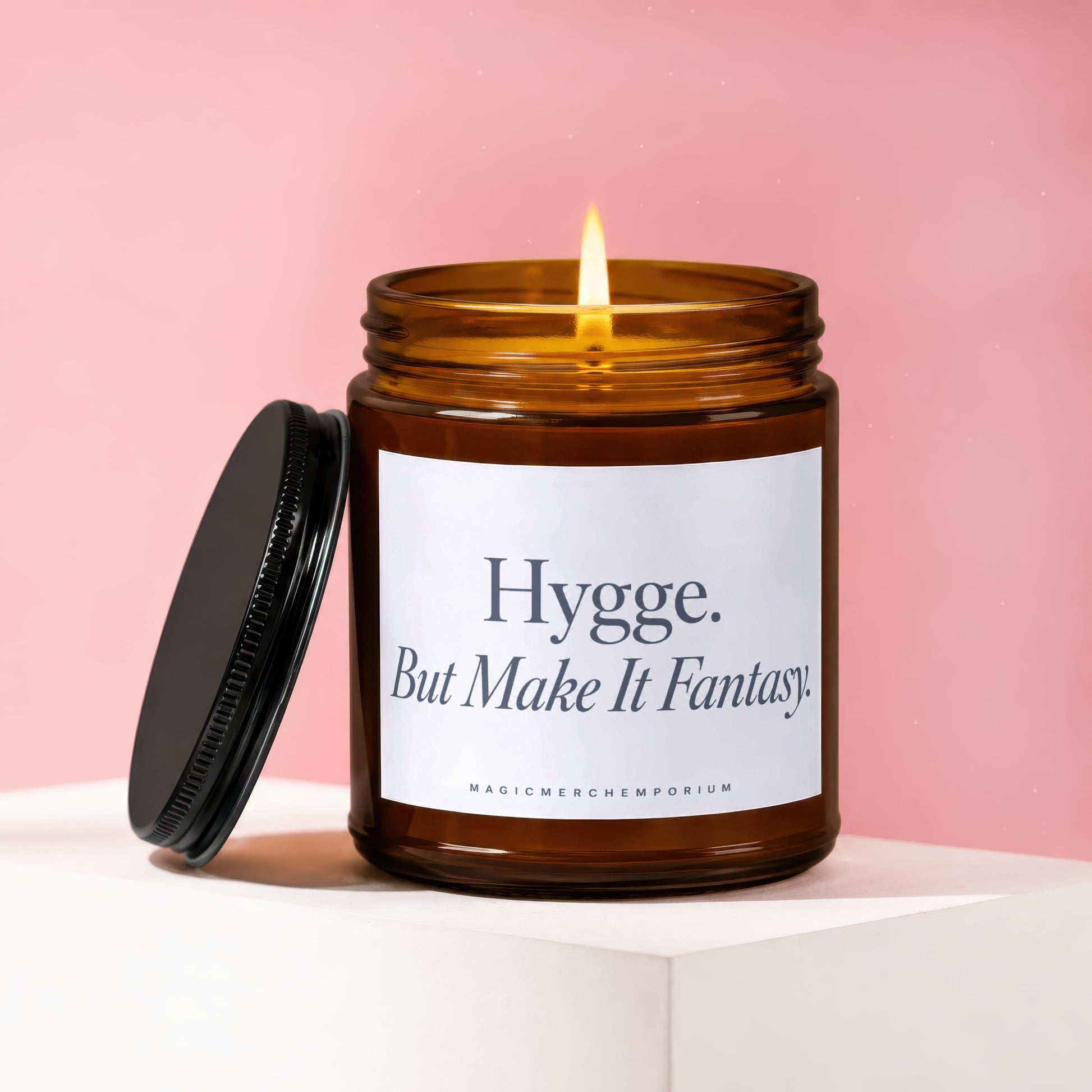 Amber jar candle with lit wick, black lid, and label reading "Hygge. But Make It Fantasy."