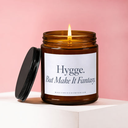 Amber jar candle with lit wick, black lid, and label reading "Hygge. But Make It Fantasy."