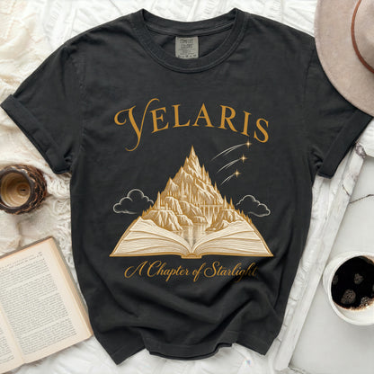 Black t-shirt with gold text "VELARIS" and mountain castle design above an open book.