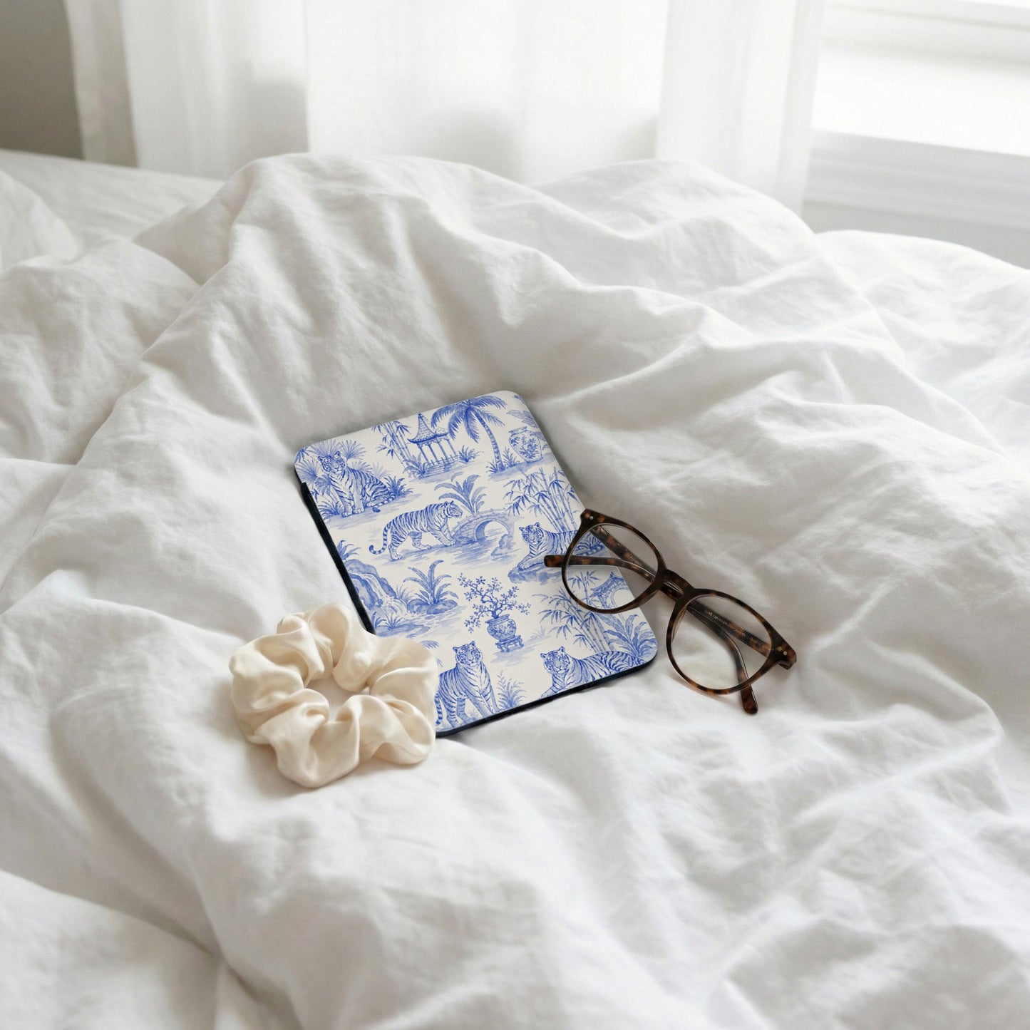 Blue and white toile pattern notebook with tigers, scrunchie, and glasses on white bedding.