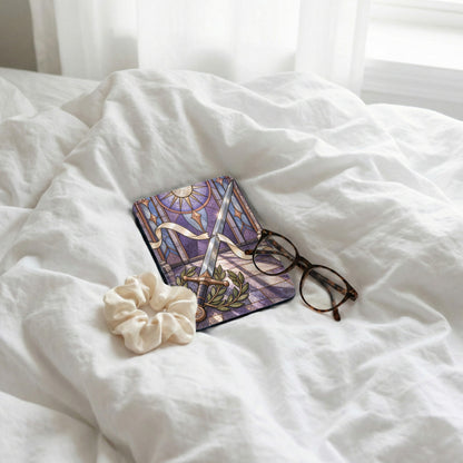 Book with stained glass sword design, scrunchie, and glasses on white bedding.