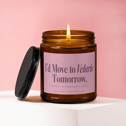 Lit amber glass candle with a pink label reading I'd Move to Velaris Tomorrow, resting on a white surface against a pink background.