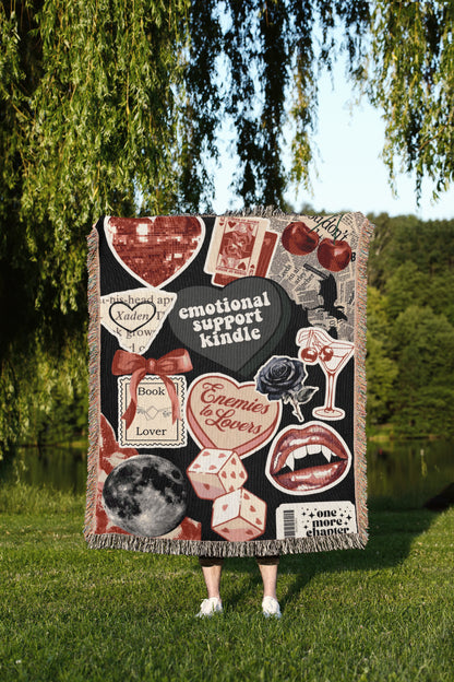 A person is standing in a grassy area, holding a large blanket with various images and text on it, including a heart, a rose, a book, and other decorative elements.