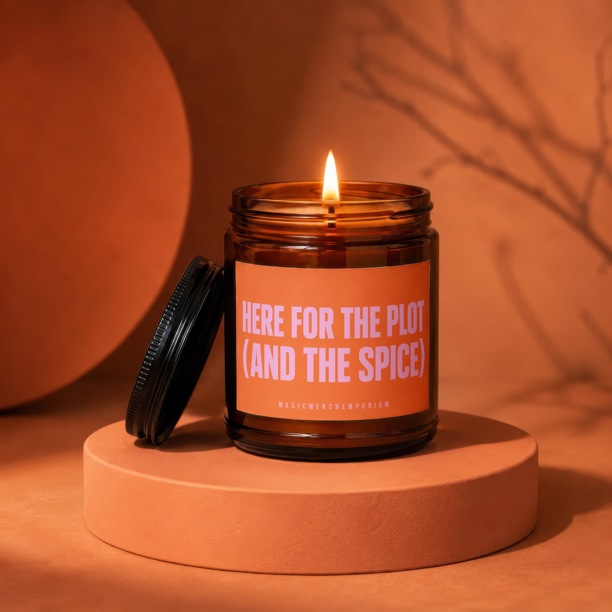 Amber jar candle with a lit wick, orange label reading "HERE FOR THE PLOT (AND THE SPICE)".