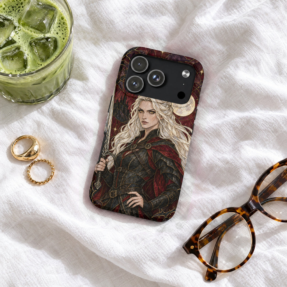 A smartphone case featuring a fantasy warrior illustration rests on white fabric next to glasses, rings, and a drink.