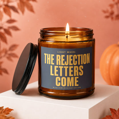 Amber candle with "The Rejection Letters Come" text, lit wick, lid off.