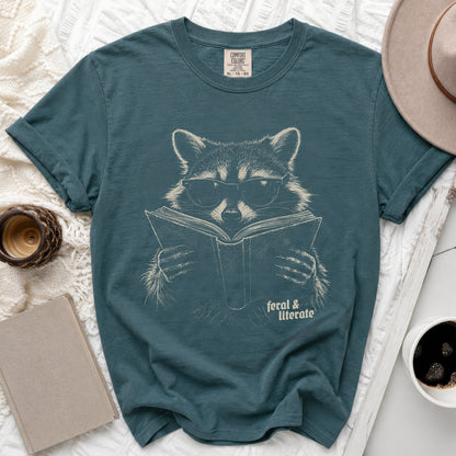 Teal t-shirt with a line drawing of a raccoon wearing glasses reading a book.