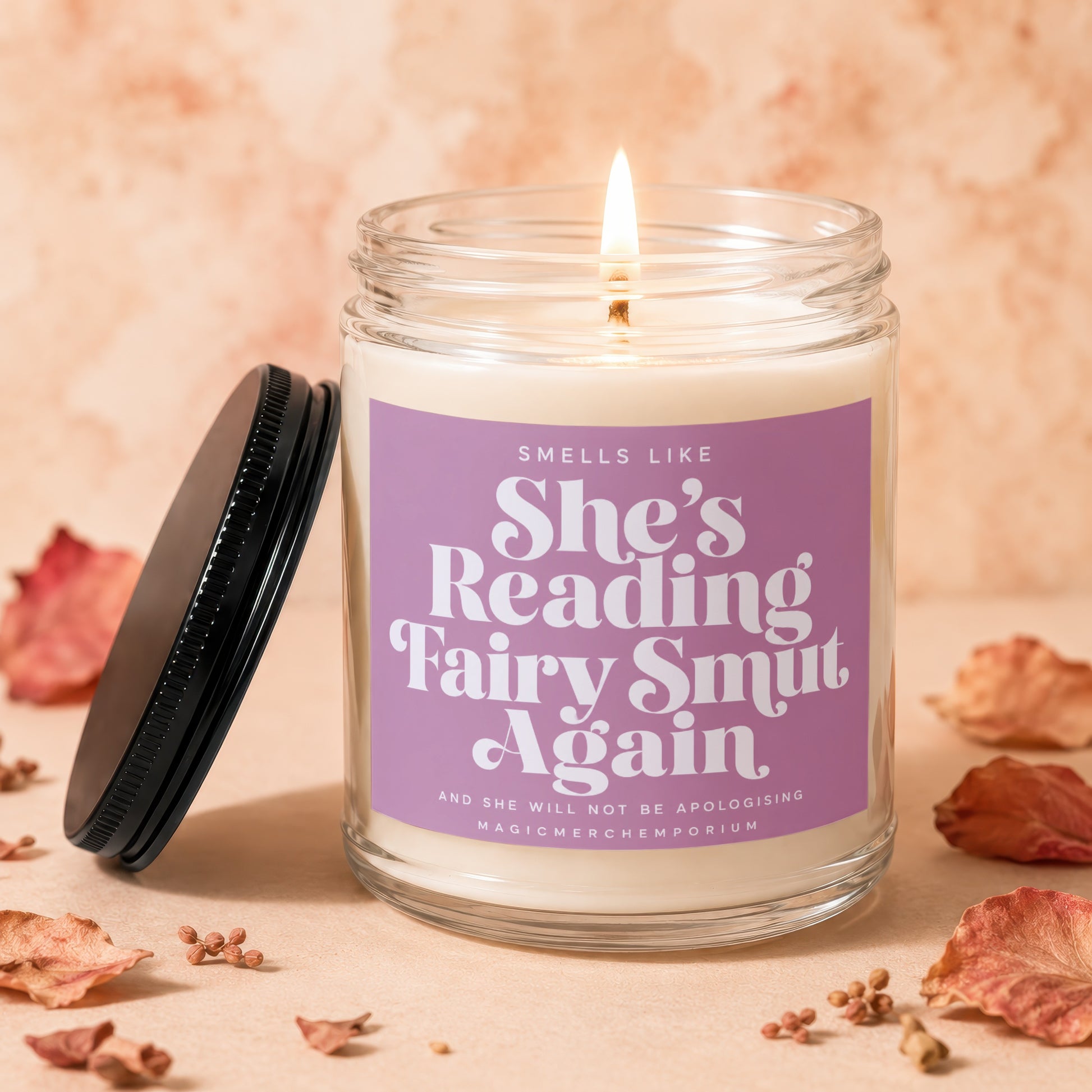 Scented candle with purple label reading "She's Reading Fairy Smut Again".