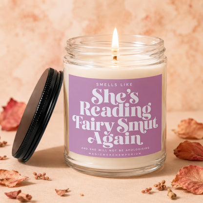 Scented candle with purple label reading "She's Reading Fairy Smut Again".