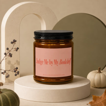 Amber candle with pink label reading "Judge Me by My Bookshelf" and "MAGICMERCHÉMPORIUM".