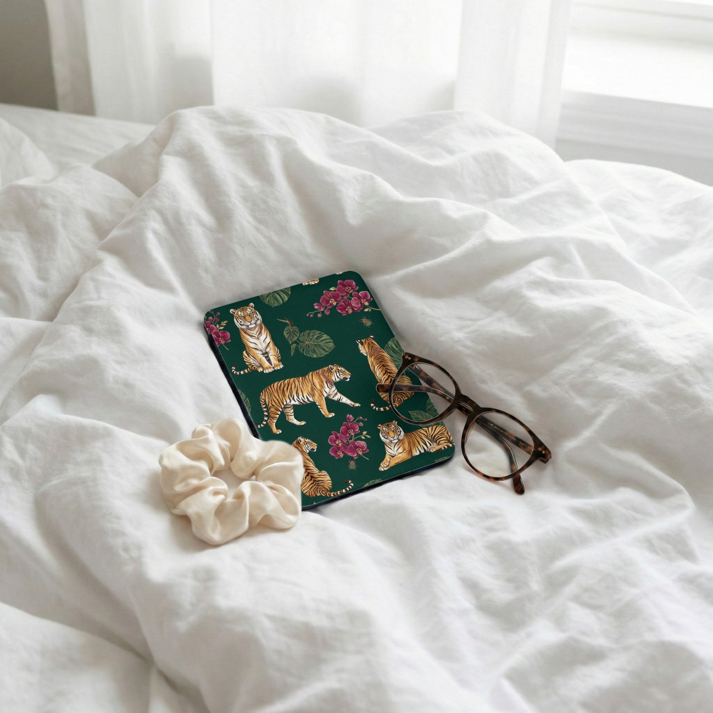 Dark green case with tigers and orchids, eyeglasses, and scrunchie on white bedding.