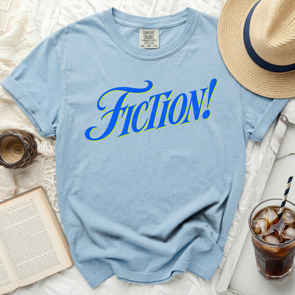 Light blue t-shirt with blue and yellow "FICTION!" graphic and Comfort Colors tag.
