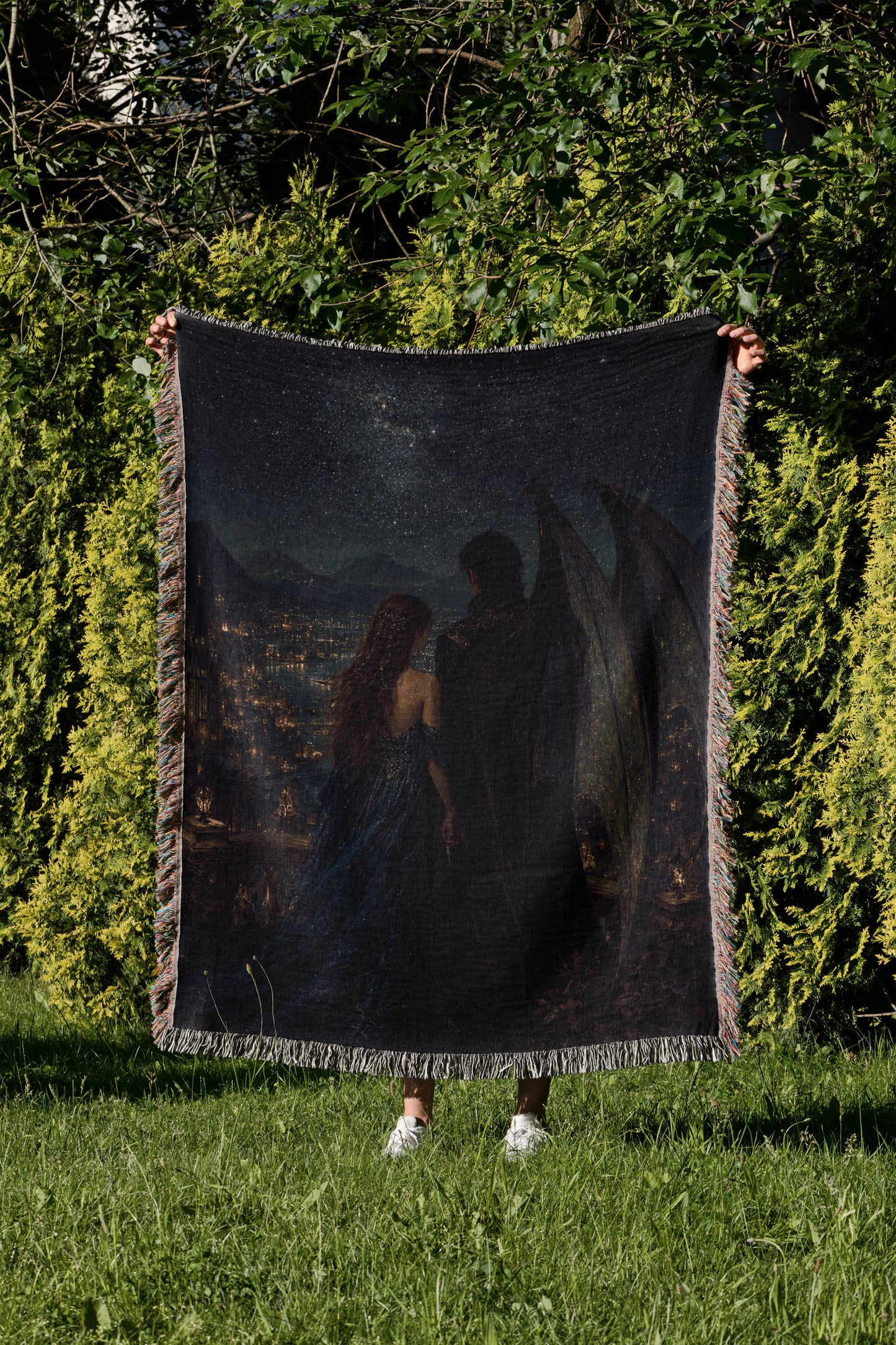 A person is standing in a grassy area, holding up a large, dark-colored blanket that features a painting of a couple.