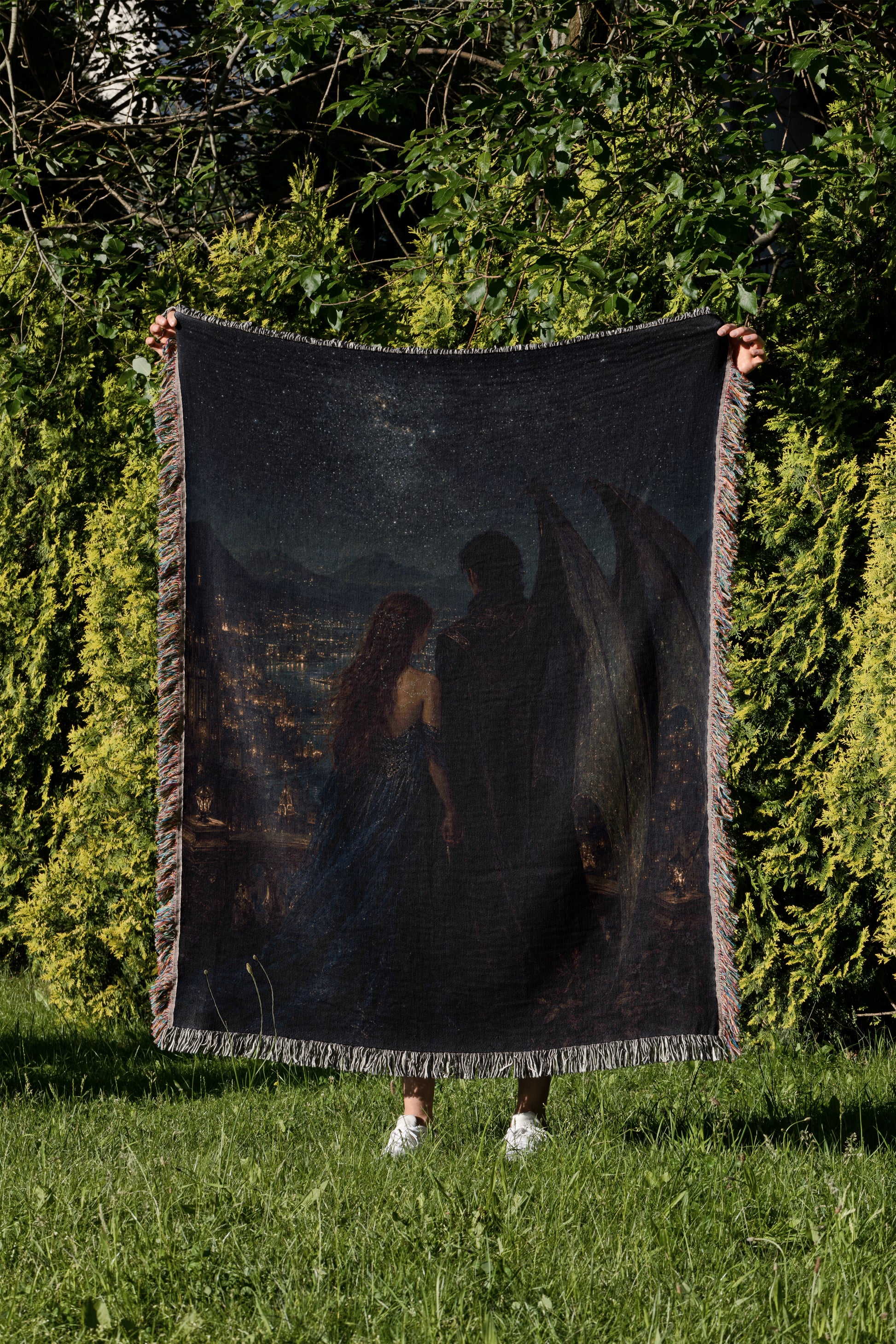 A person is standing in a grassy area, holding up a large, dark-colored blanket that features a painting of a couple.