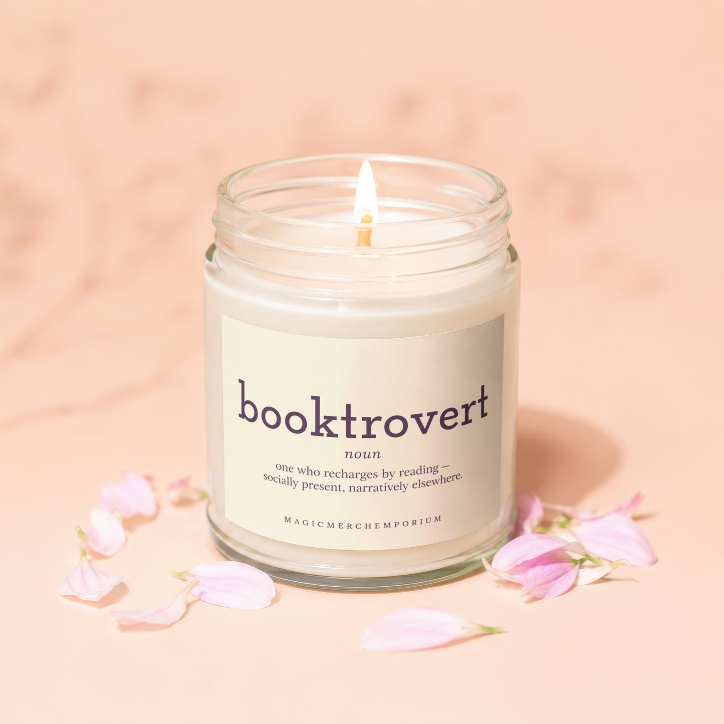 Glass jar candle with "booktrovert" definition and pink petals on a light peach background.