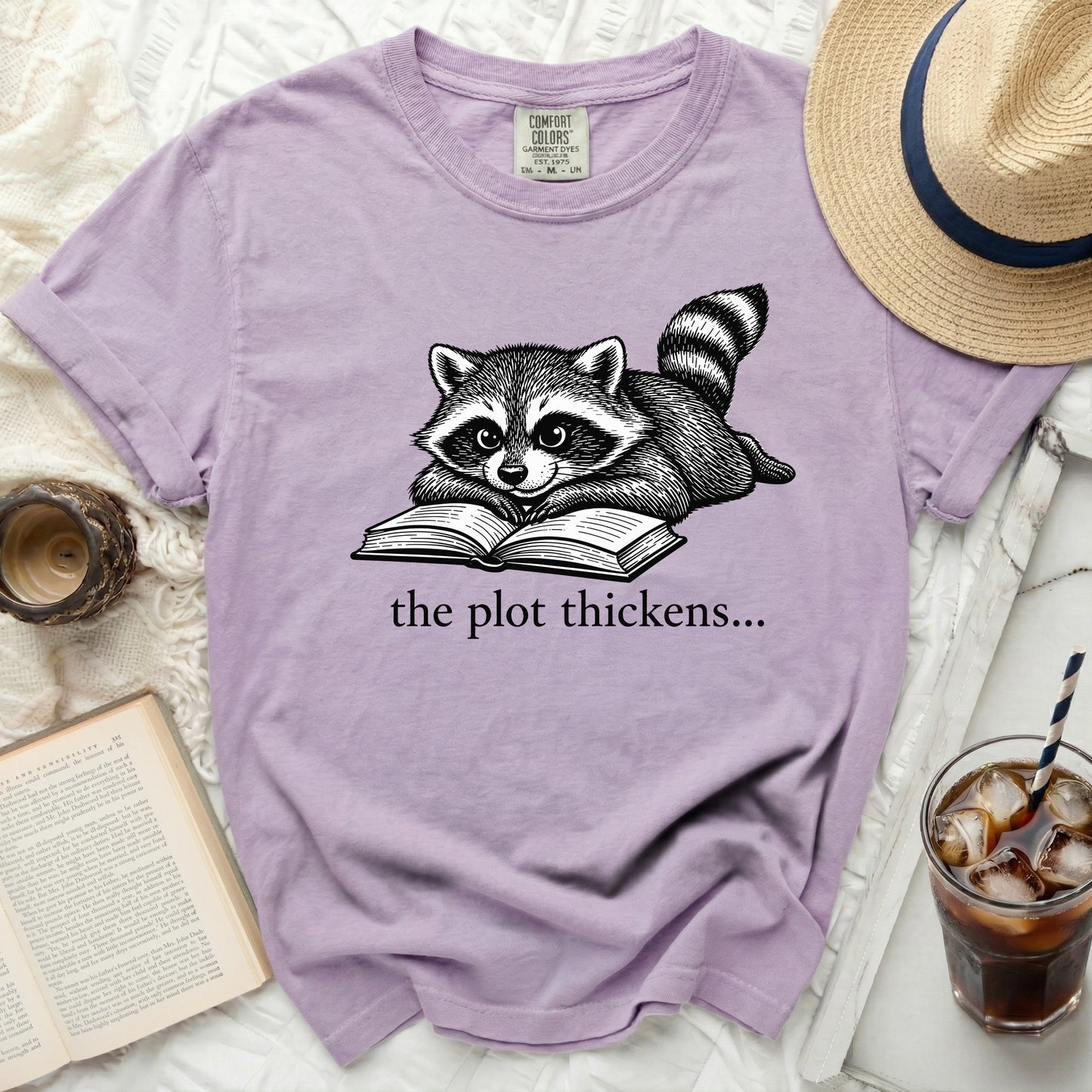 Purple t-shirt with a raccoon reading a book graphic and text "the plot thickens..."