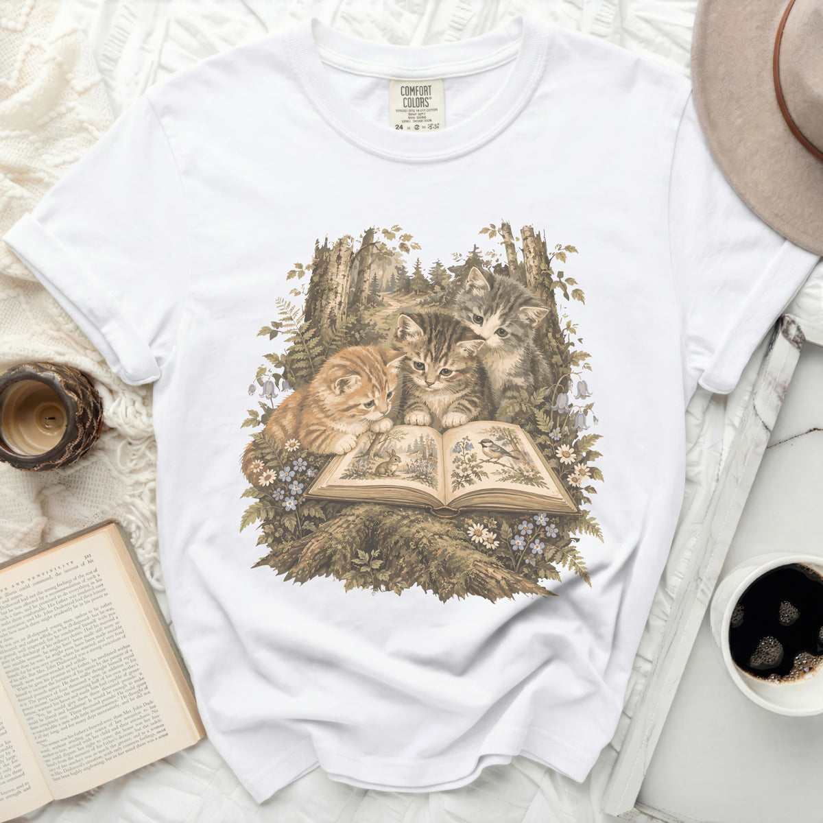 Three kittens gathered around an open book in a forest setting on a white t-shirt.