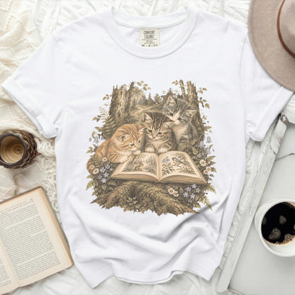 Three kittens gathered around an open book in a forest setting on a white t-shirt.
