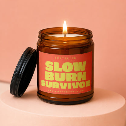 Amber glass candle with lit wick, featuring "SLOW BURN SURVIVOR" on an orange label.