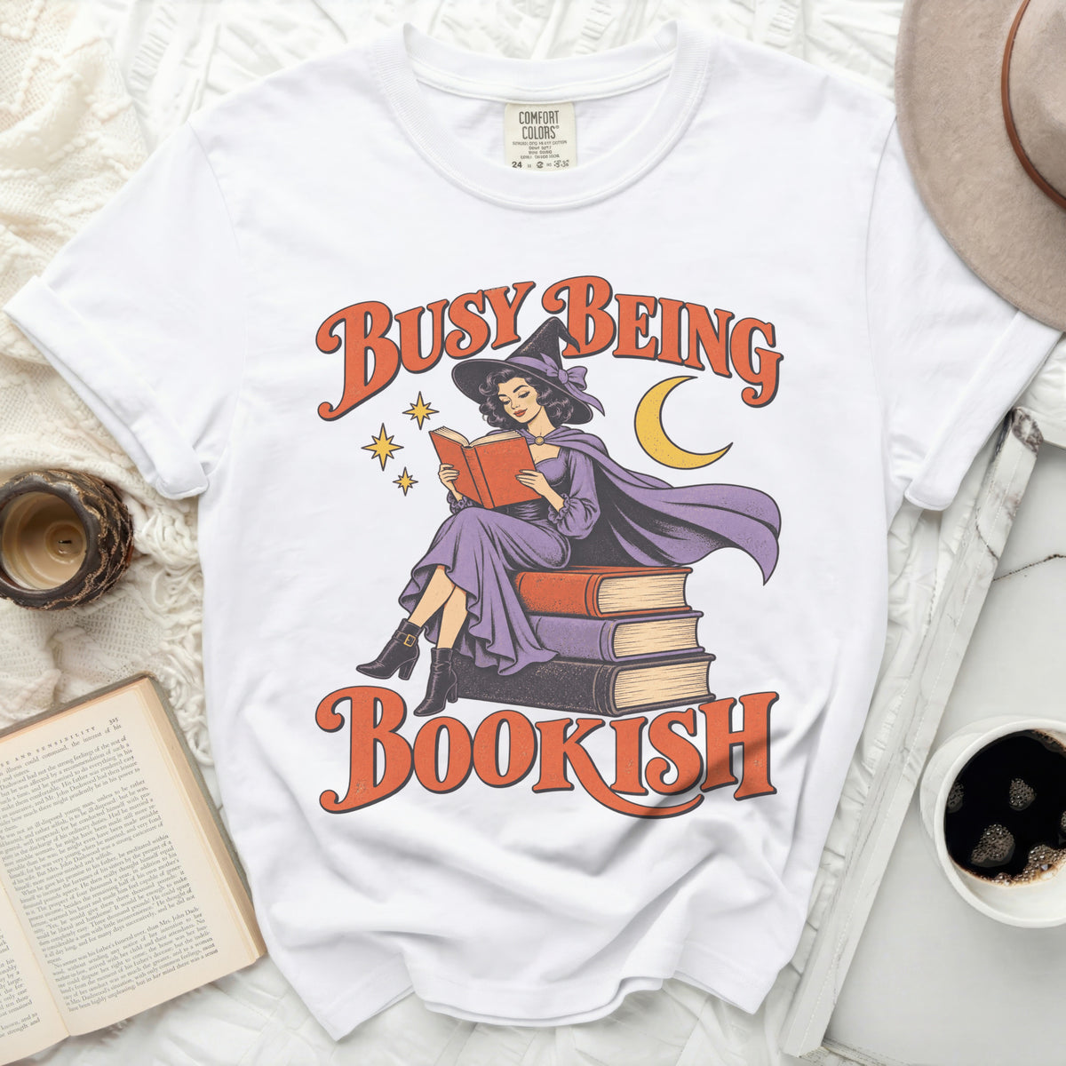 White t-shirt with "Busy Being Bookish" graphic of a witch reading.