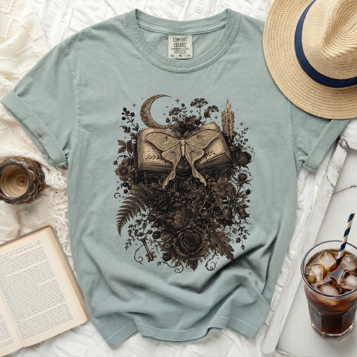 Light blue t-shirt with moth, book, moon and floral graphic design.