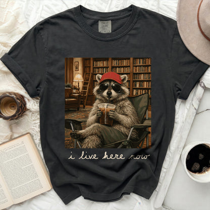 Raccoon wearing sunglasses and a hat drinks iced coffee in a library, text says "i live here now".