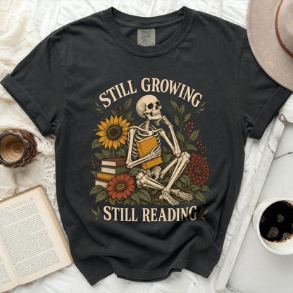 Dark t-shirt with skeleton, books, flowers, and text "Still Growing Still Reading".