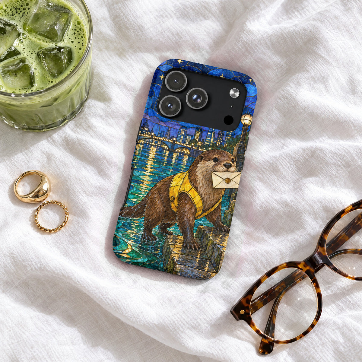 A phone case with a stained-glass style otter carrying a letter rests on white fabric next to glasses, rings, and iced green tea.