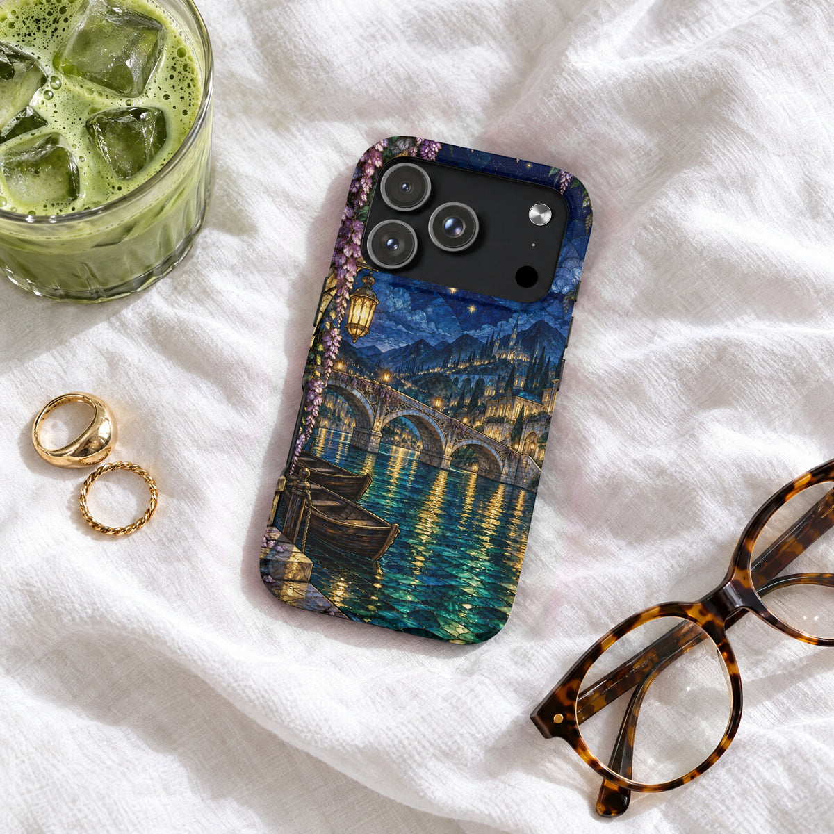 Blue phone case featuring a moonlit Venetian canal scene with stone bridges, surrounded by glasses, rings, and an iced drink.
