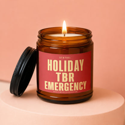 Amber jar candle with lit wick and red label reading "STATUS: HOLIDAY TBR EMERGENCY MAGICMERCHEMPORIUM".