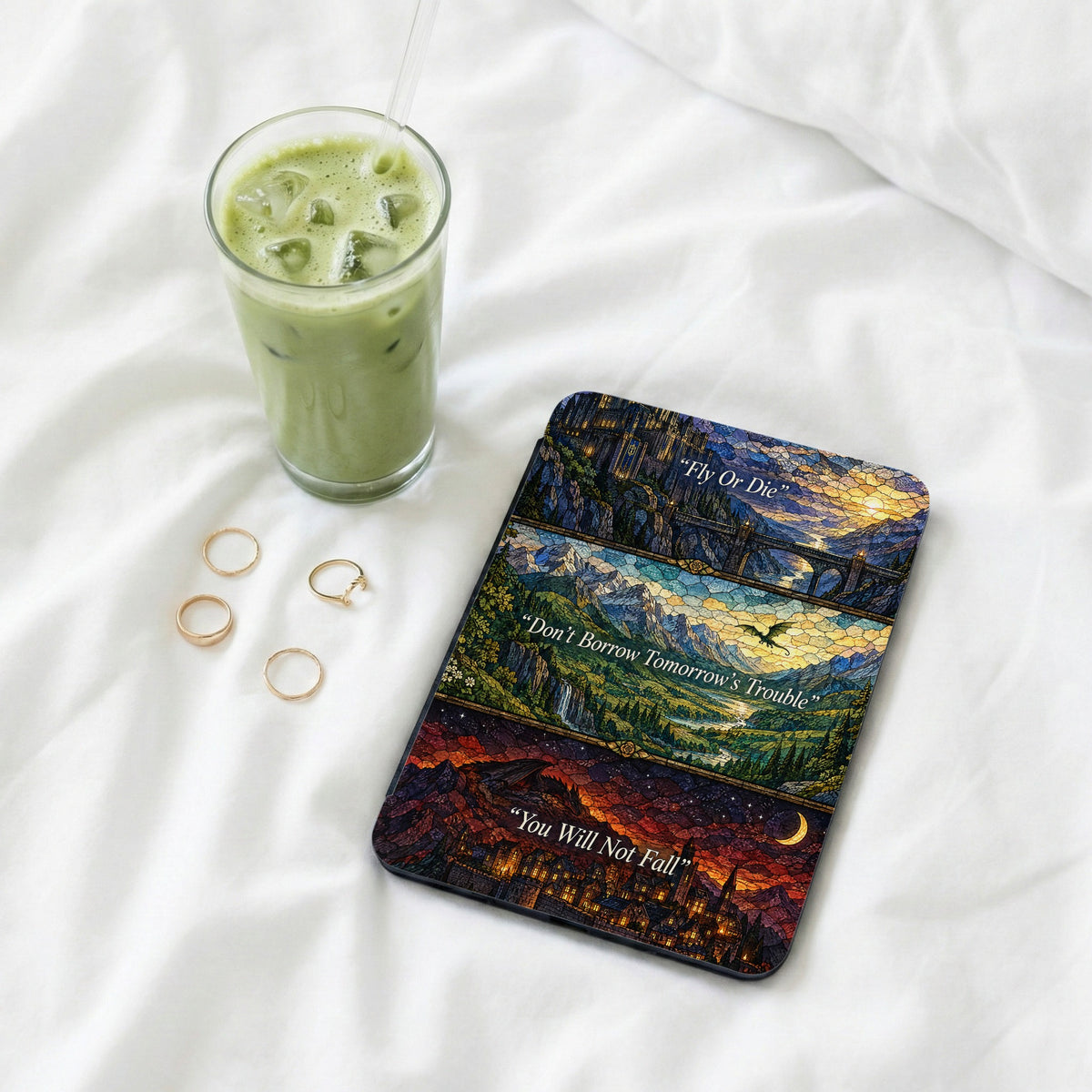 A Kindle case with stained glass art rests on a white bed with rings and a matcha latte.