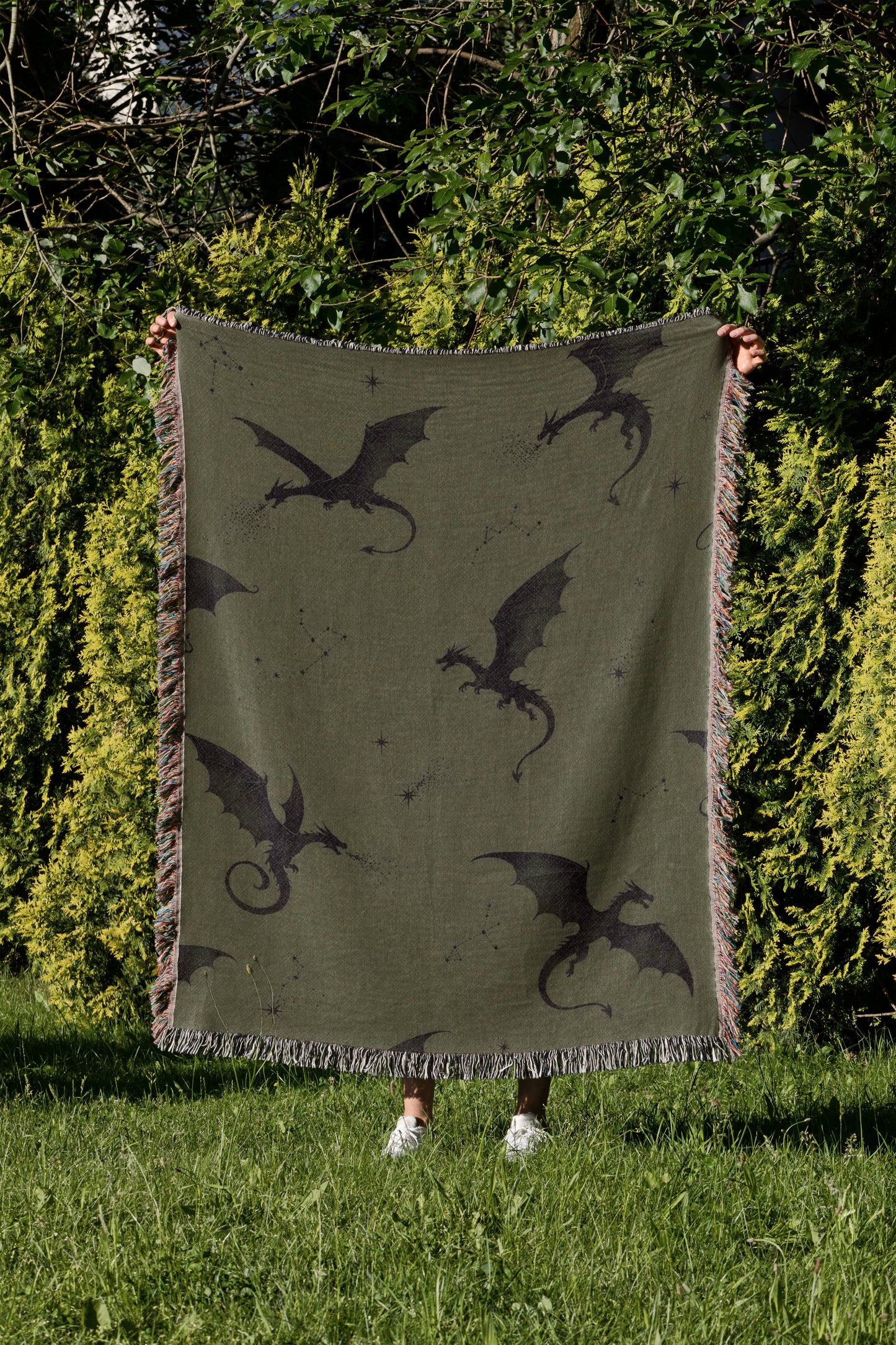A person is holding up a large green blanket with black dragon silhouettes on it, standing in a grassy area with trees in the background.
