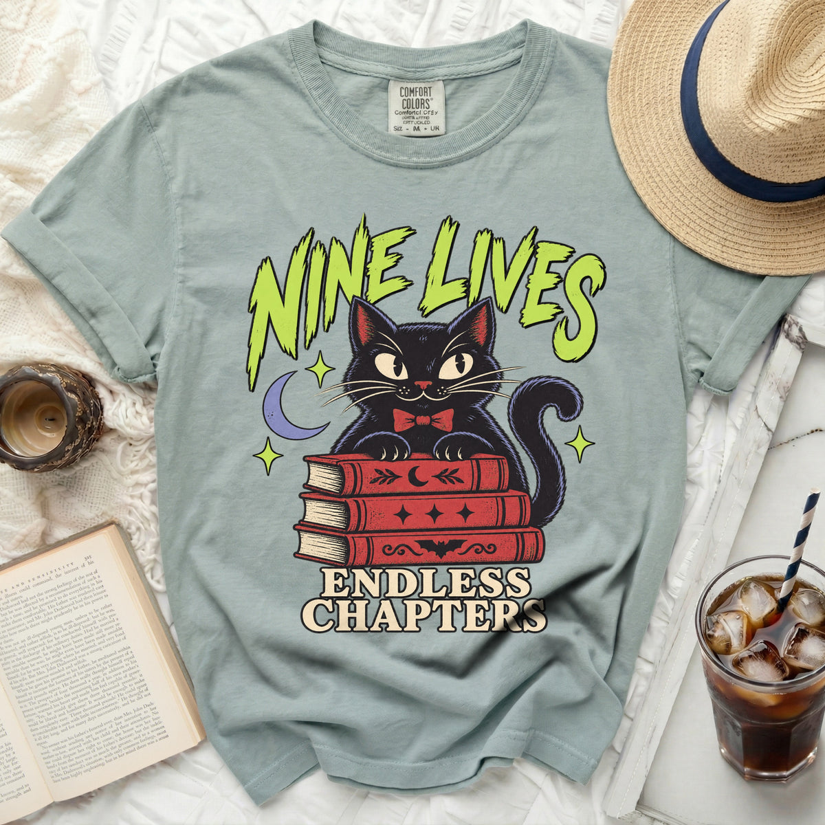 Green t-shirt with black cat on books reading "Nine Lives Endless Chapters."