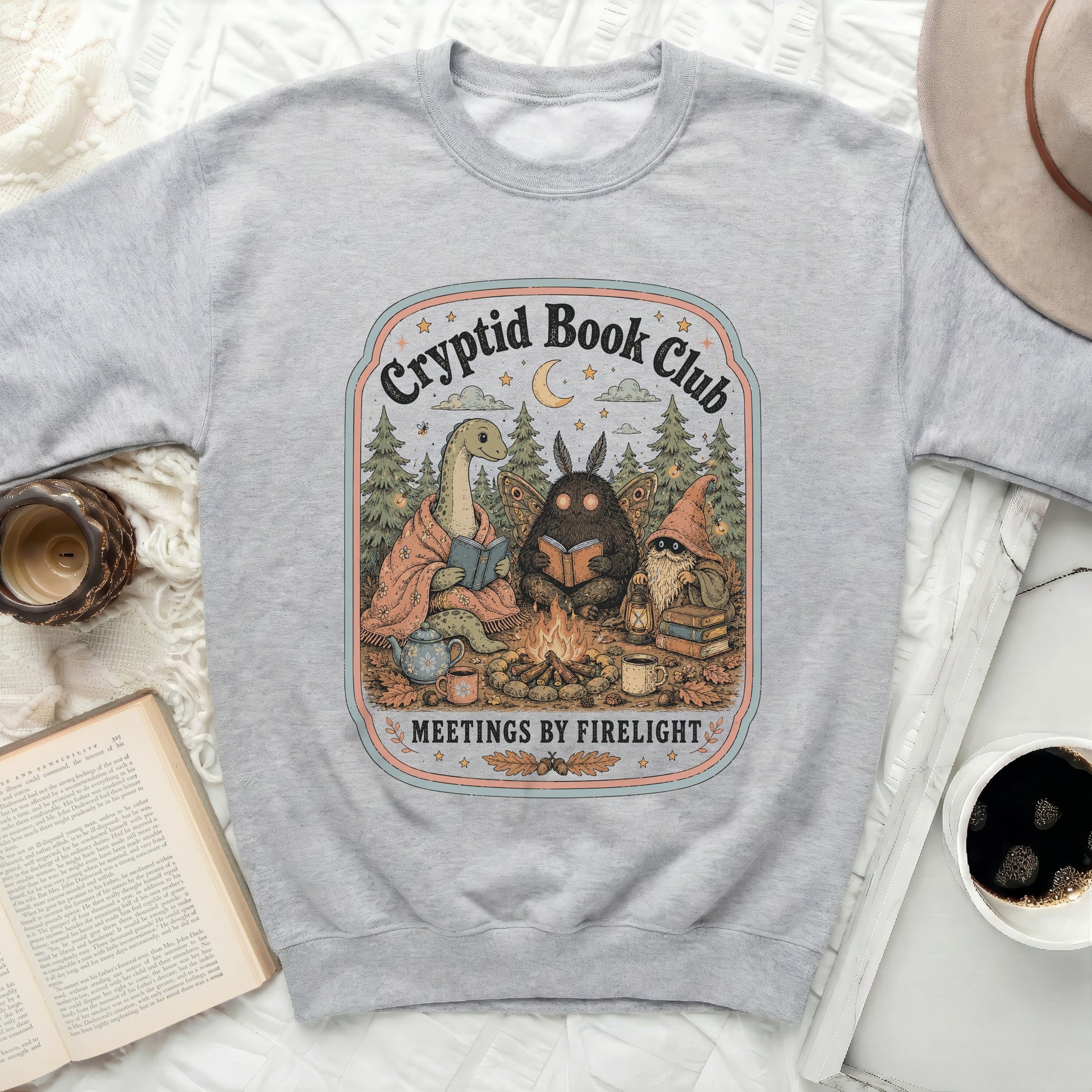 Gray sweatshirt with "Cryptid Book Club Meetings by Firelight" illustration.