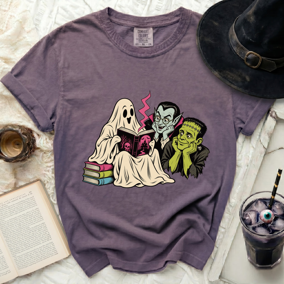 Ghost, vampire, and Frankenstein read a spooky book on a purple t-shirt.